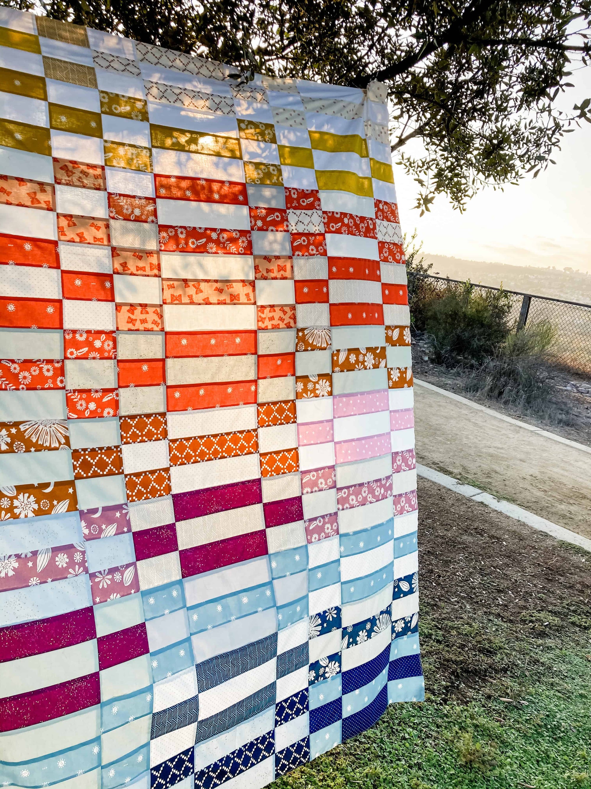 a rainbow patchwork quilt in rectangular grid pattern hanging near a tree at sunset