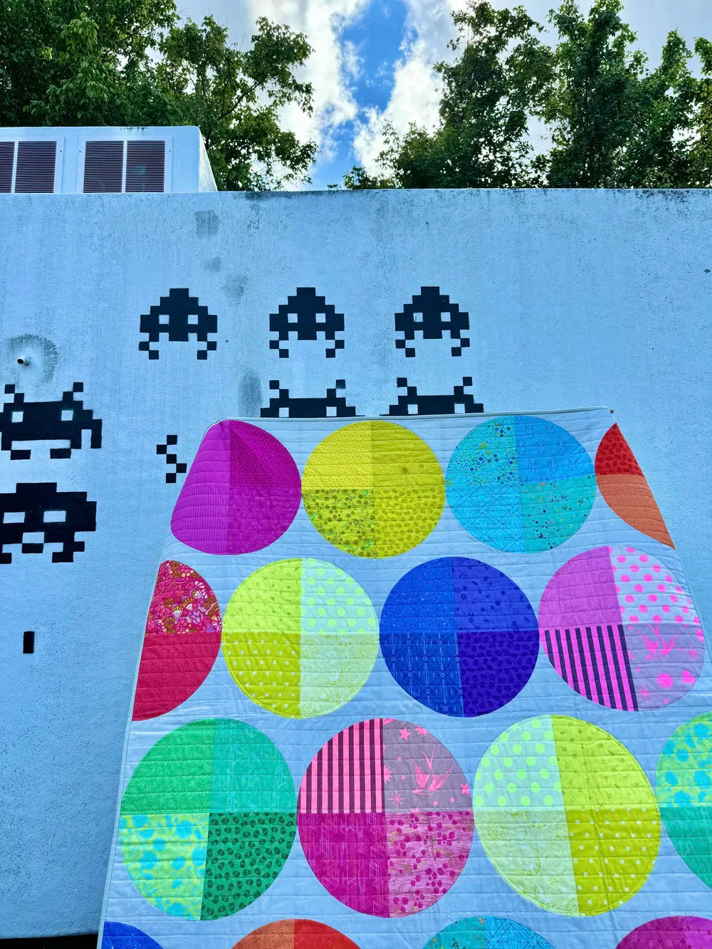 patchwork quilt in neon fabrics in circle shapes, held in front of a space invaders mural