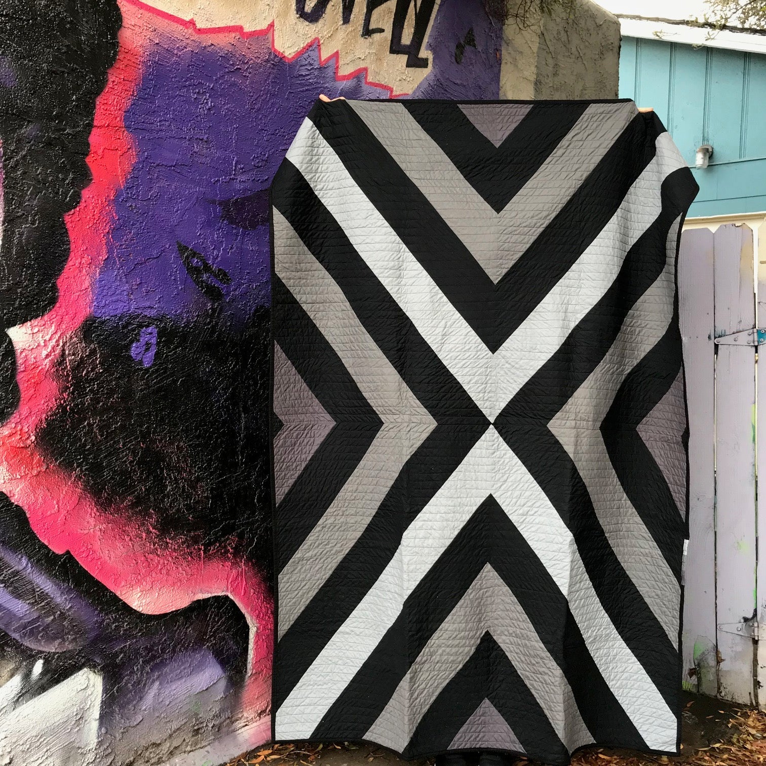 minimalist quilt with bold patchwork X in grays and black in front of a colorful mural wall