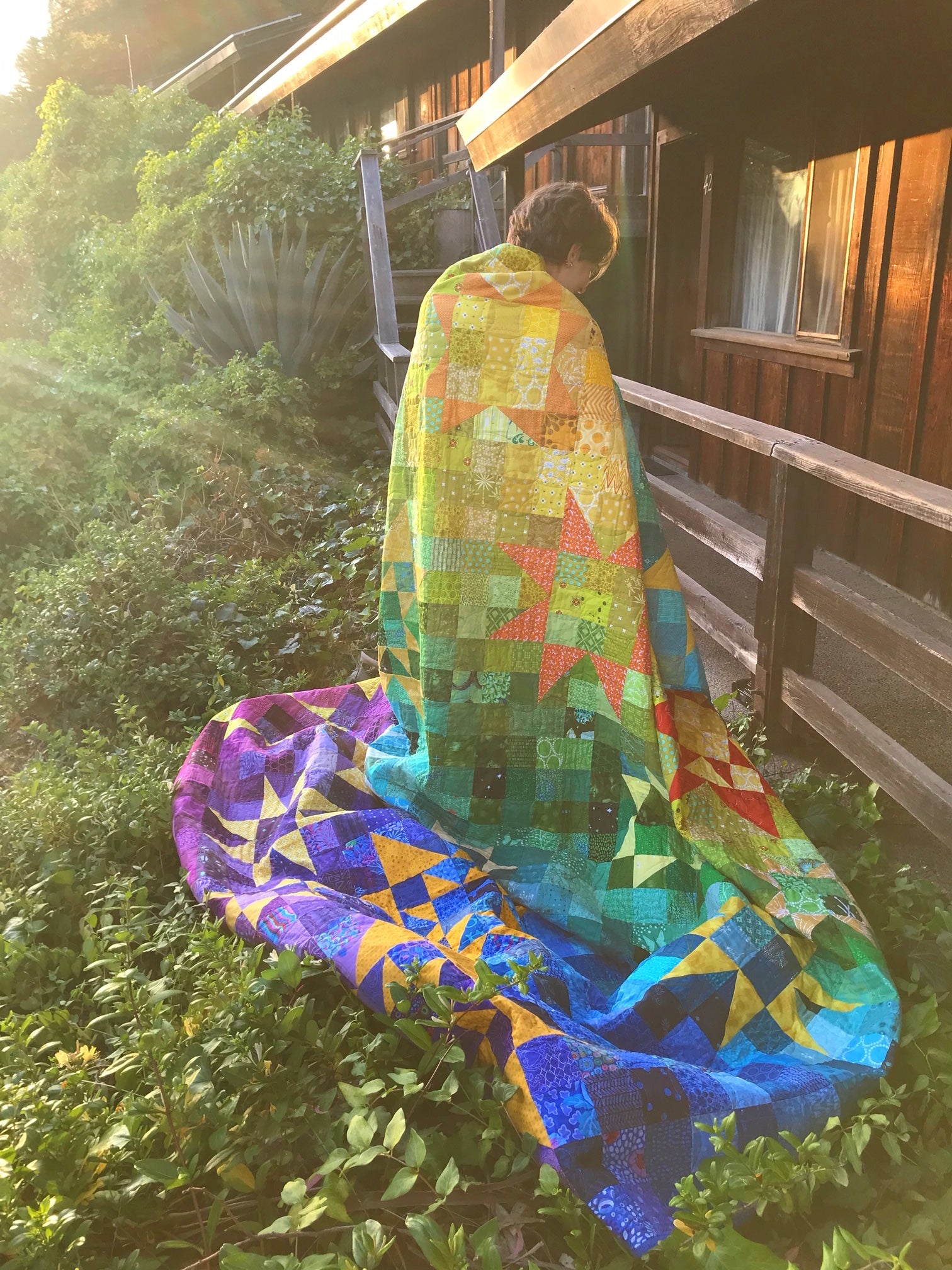 person standing in sunbeam wrapped in huge rainbow gradient patchwork quilt with stars.