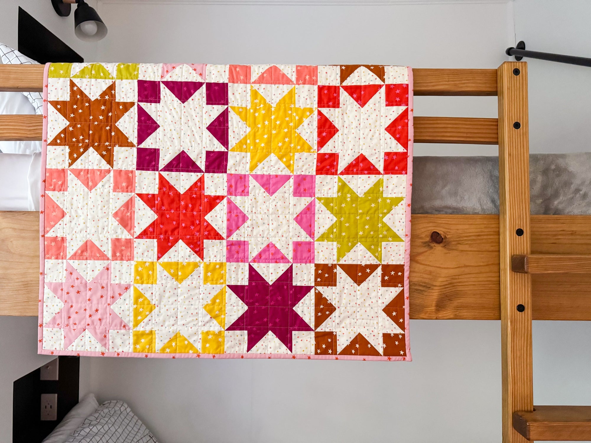 Colorful baby quilt with sawtooth star blocks in pink, red, orange, and yellow on a white background, displayed on a wooden quilt ladder.