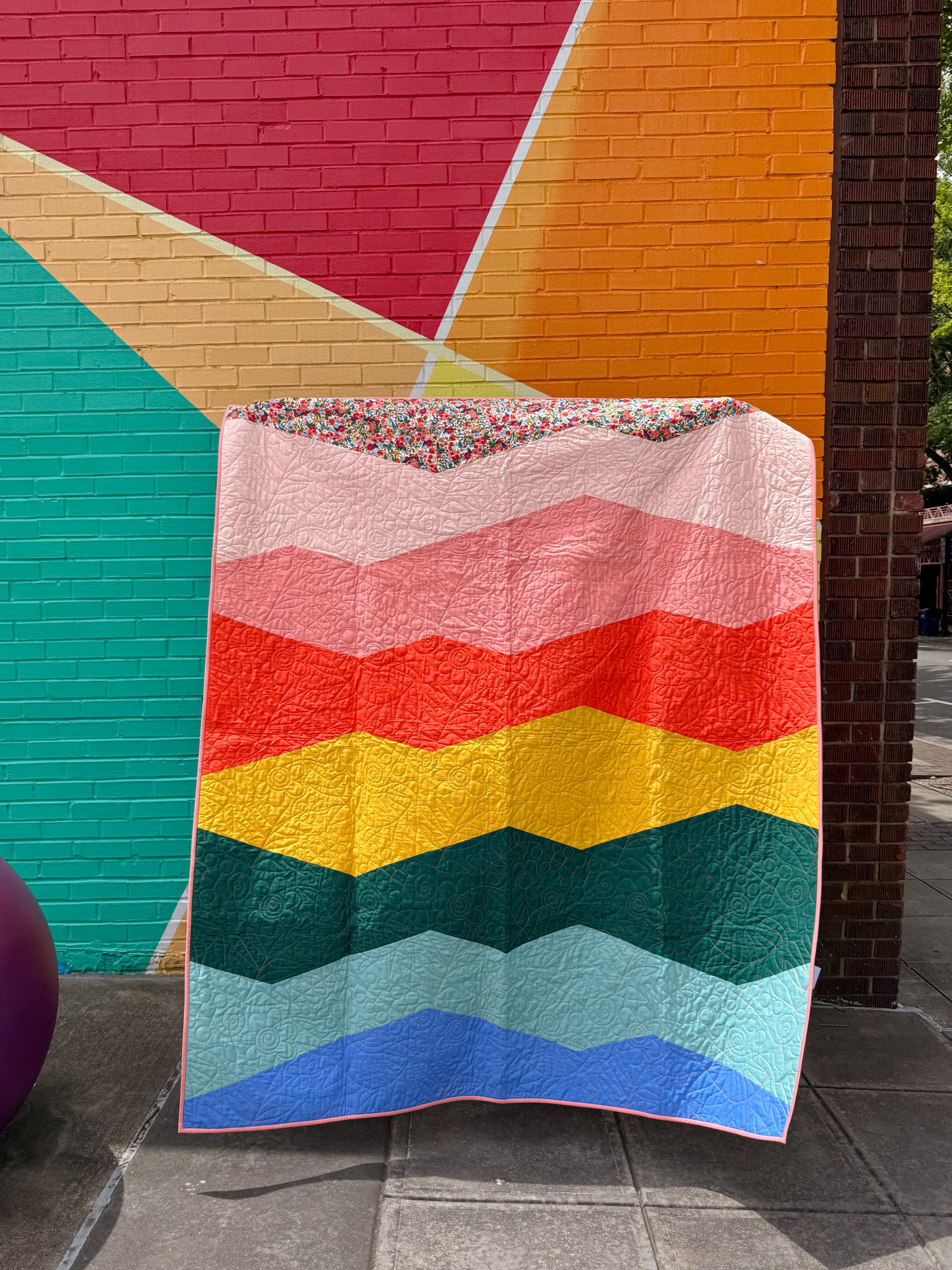 Colorful modern patchwork quilt with wavy geometric pattern draped against a multicolored wall.