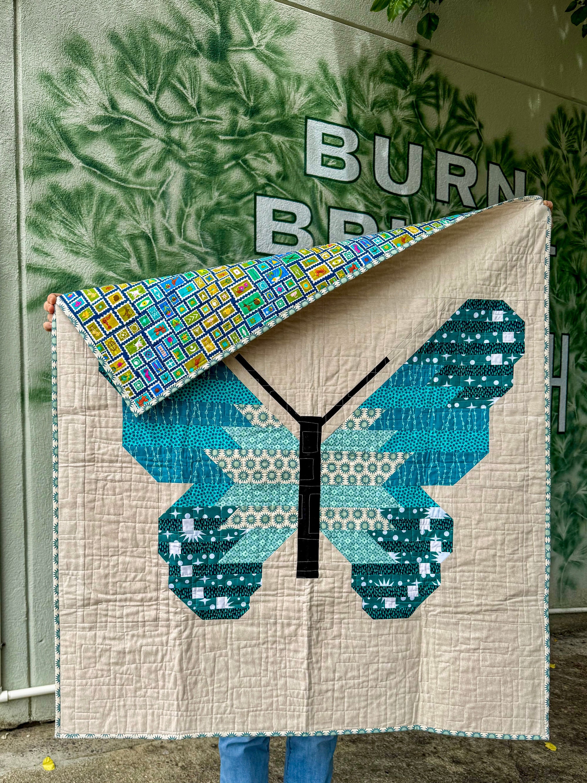 Person holding a quilt with a butterfly design in front of a building with plant painting on the wall