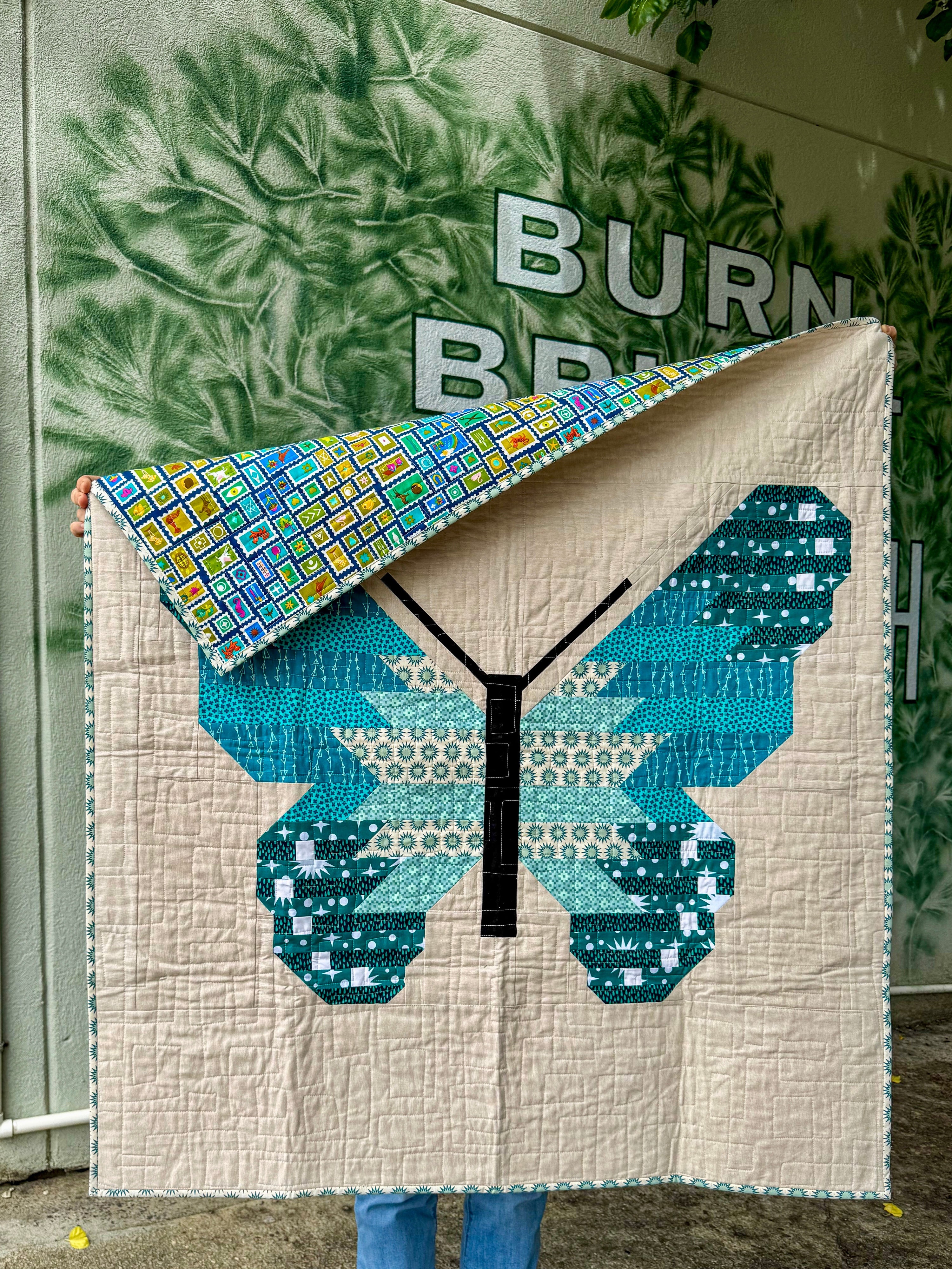 Person holding a quilt with a butterfly design in front of a building with plant painting on the wall
